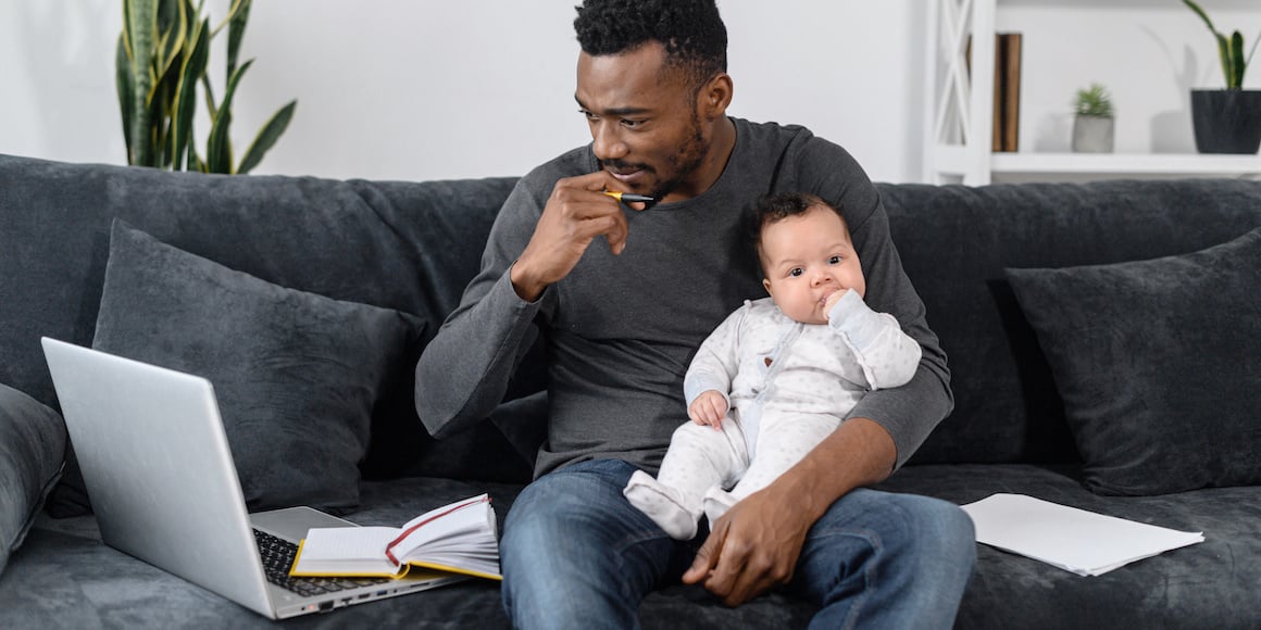 A thoughtful online, student dad with an infant sitting on the couch, he completes his MVNU Online course work from home.