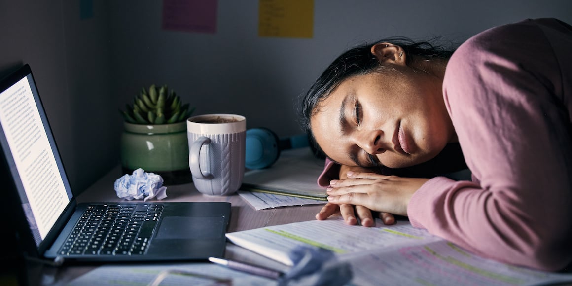 College student asleep at laptop on desk surrounded by papers and schoolwork